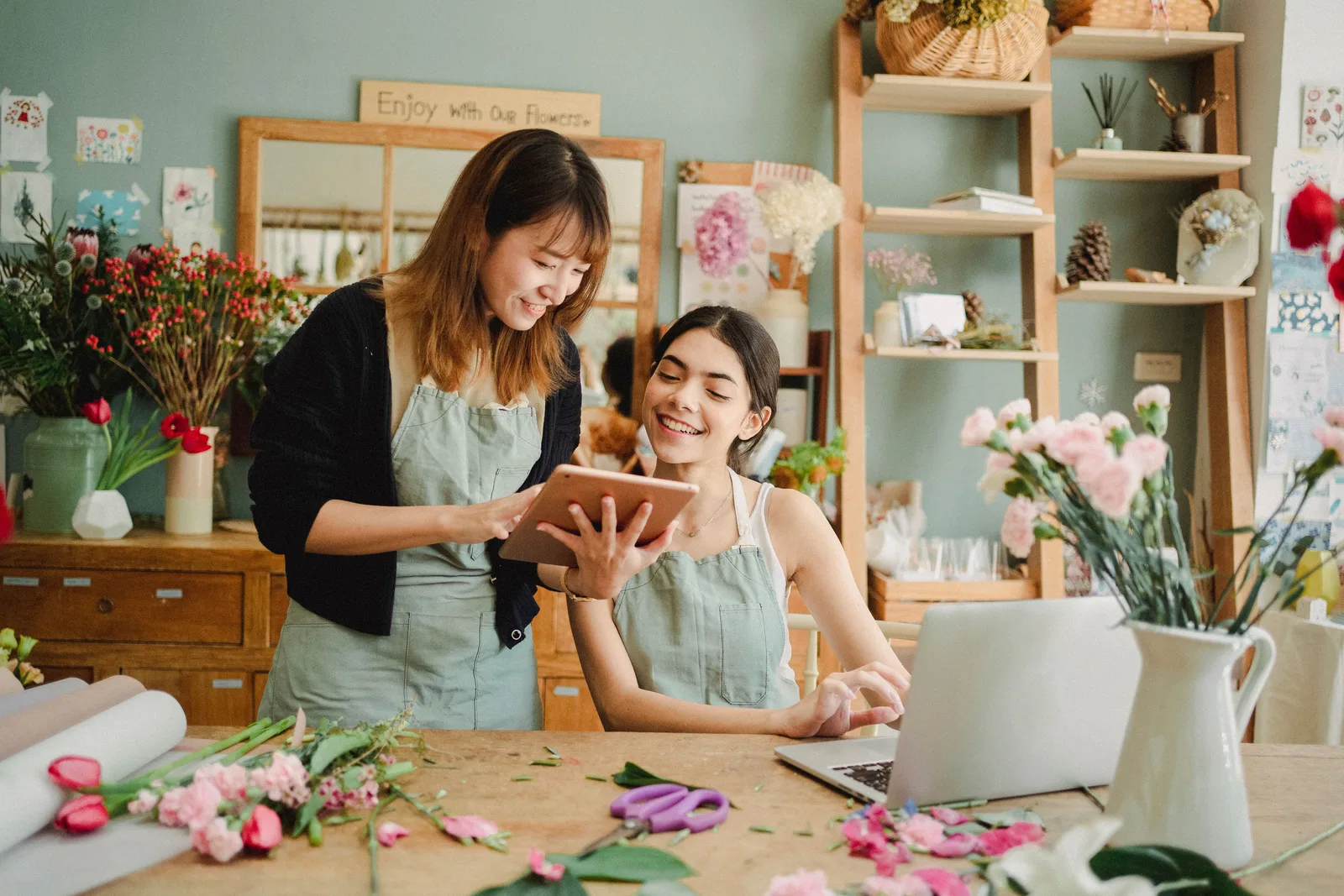 Content multiethnic female florists in aprons at table with laptop and browsing tablet while preparing for order in creative floristry studio. A local SEO checklist florists can actually run before ranking issues start